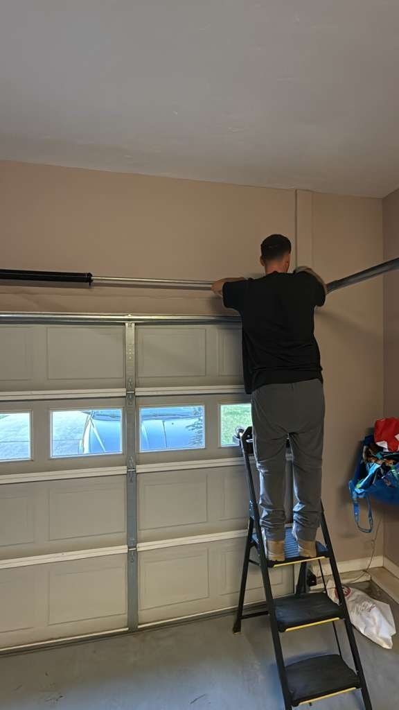 Worker on a stepladder at the top section of a windowed garage door, seen from inside