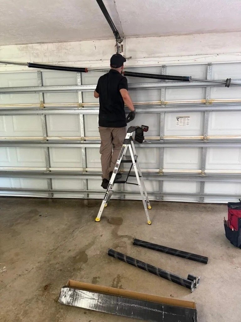 Technician on a stepladder at the spring tube above a garage door, with parts laid out on the floor