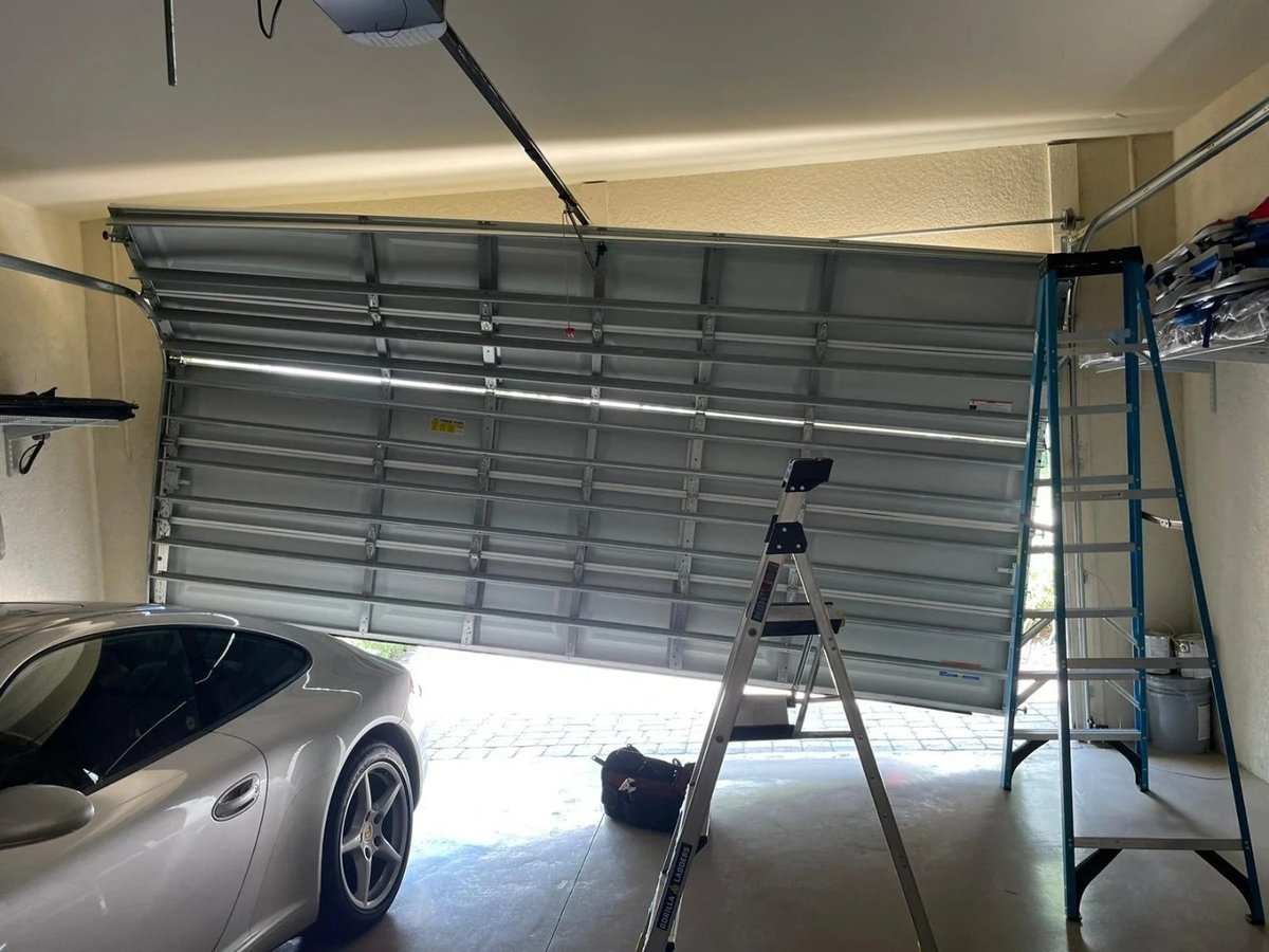 Tilted sectional garage door with ladders propped under it and a car parked inside