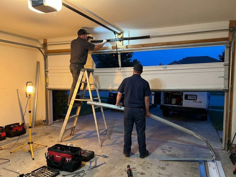 Two technicians at dusk working beside a ladder and a raised garage door under work lights